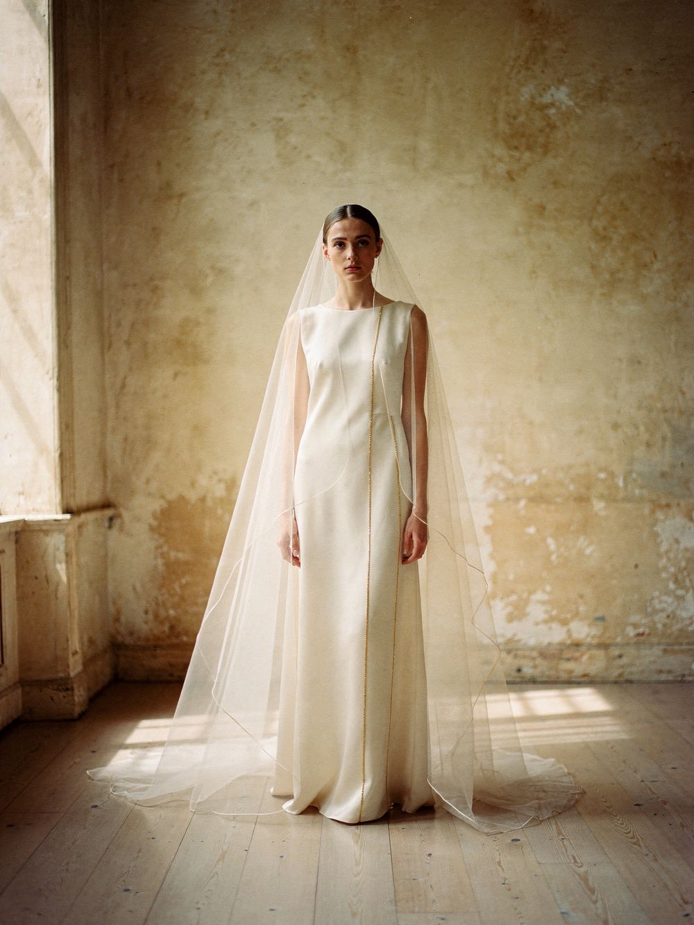 Look 01, Le Voile: an ivory silk column gown with gold-thread seams beneath a floor-length sheer veil, in a sunlit atelier