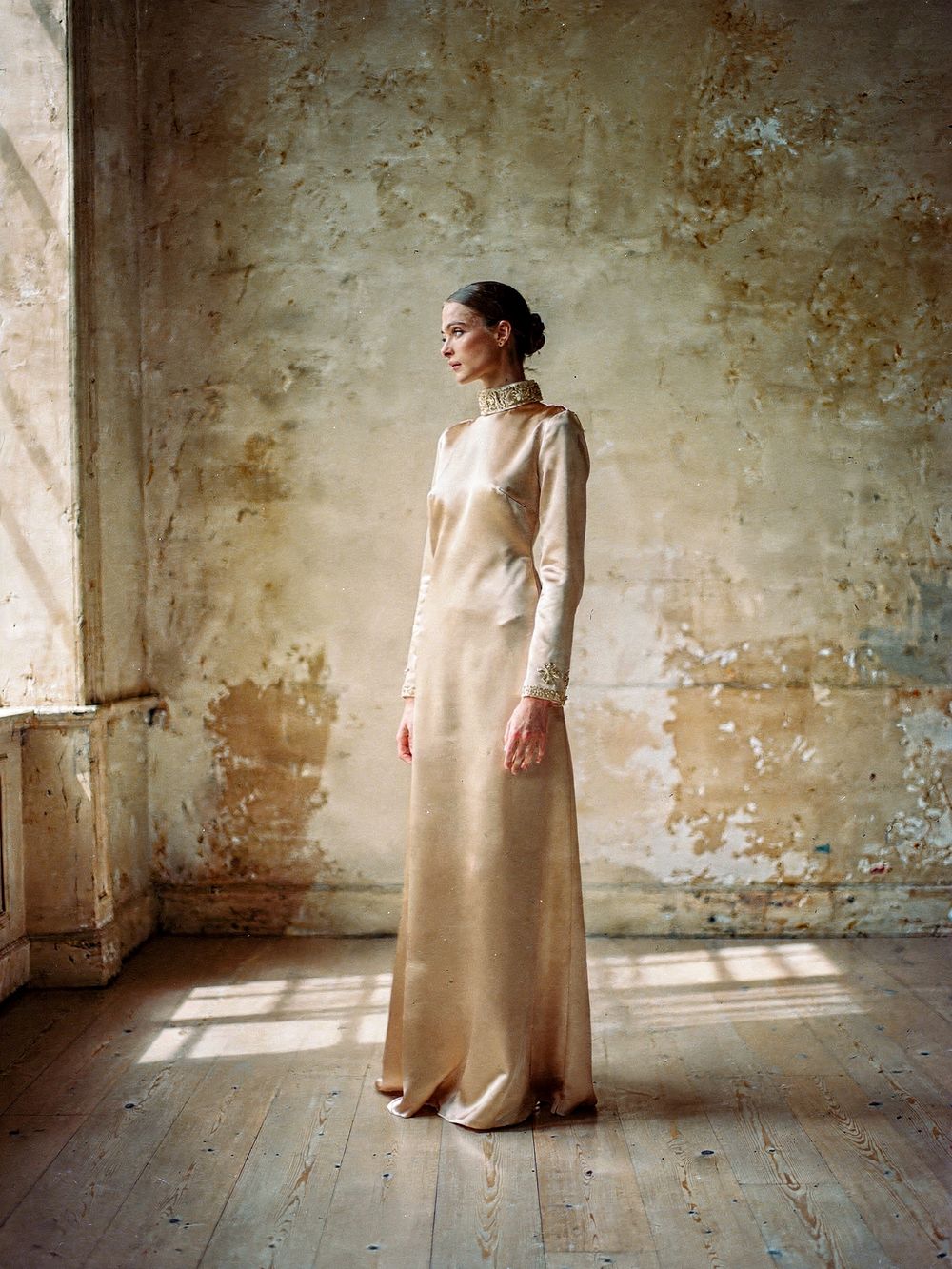 Look 02, L'Or: a long-sleeved champagne satin gown with gold embroidery at the high collar and cuffs, turned toward the window